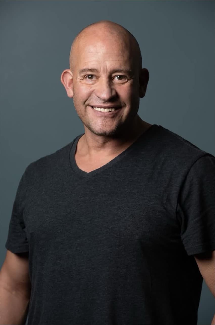 Keith Banner, a bald man in a charcoal V-neck T-shirt, smiling at the camera against a grey studio backdrop.
